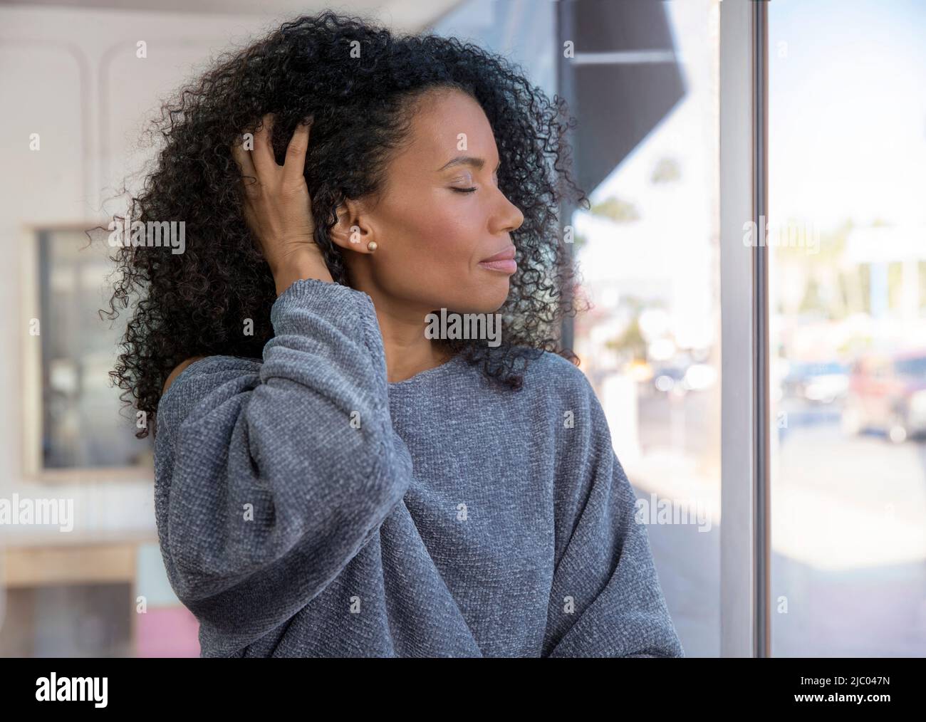 Razza mista, donna di mezza età corre le mani attraverso i capelli con gli occhi chiusi guardando fuori una finestra. Foto Stock