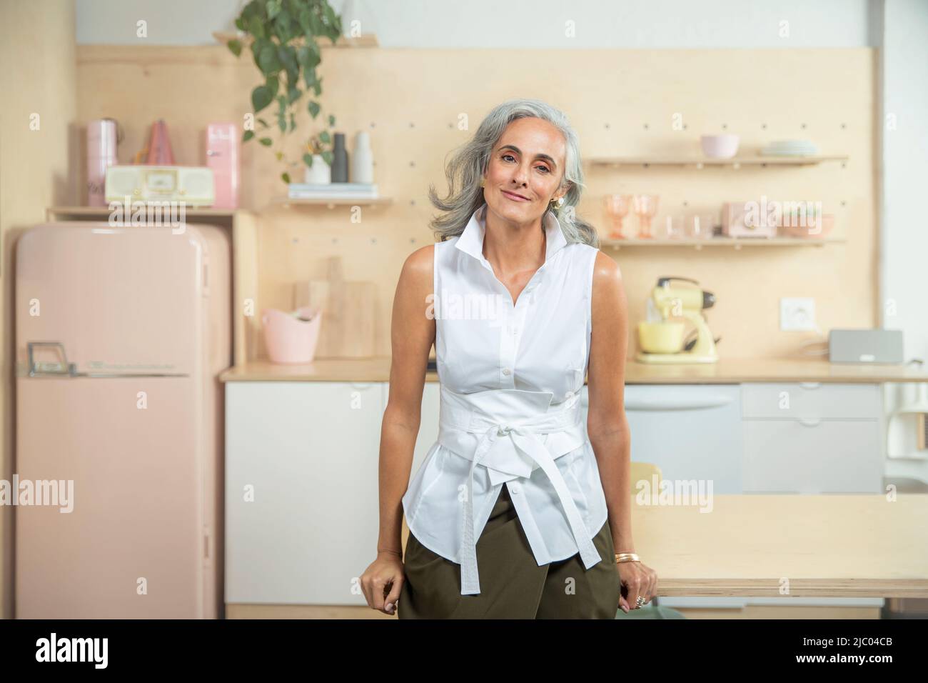 Ampio ritratto orizzontale di una donna di mezza età con capelli grigi, in piedi in una cucina guardando in macchina fotografica con sorriso sincero. Foto Stock