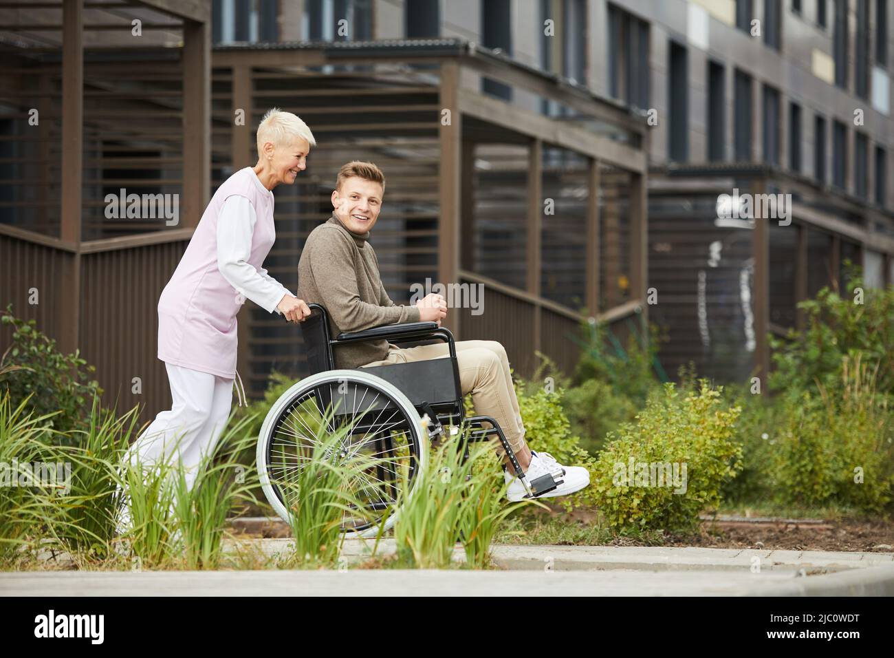 Una donna energica e positiva in uniforme medica che spinge la sedia a rotelle con il ragazzo mentre camminano in città Foto Stock