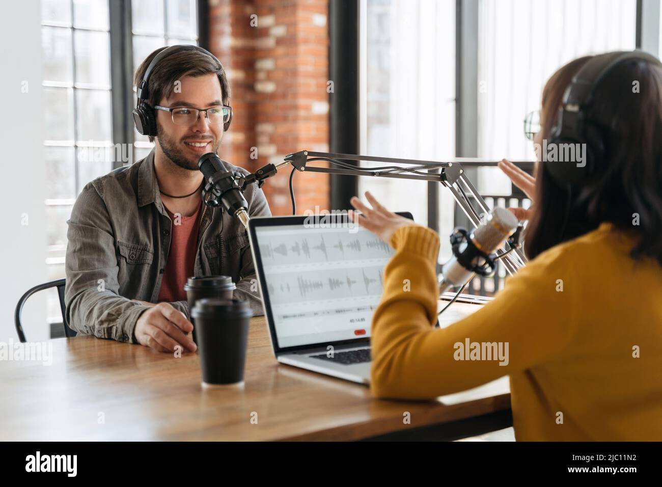 Sorridente bell'uomo che parla con gesturing bella donna radio ospite mentre registrano live audio podcast in studio. Due persone che utilizzano computer portatile, cuffie e apparecchiature audio per la realizzazione di podcast Foto Stock