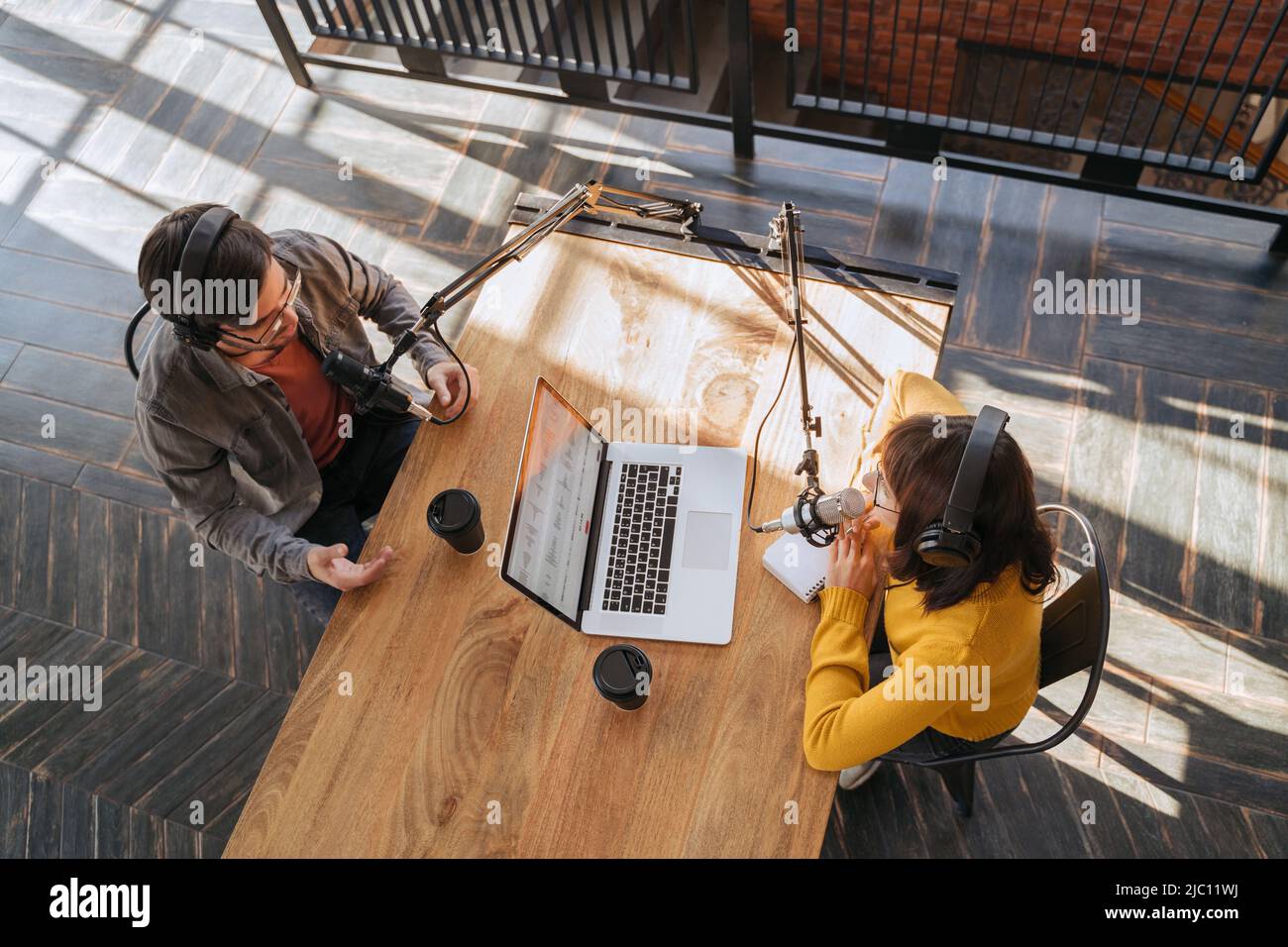 Vista dall'alto di due persone in cuffie che registrano podcast audio utilizzando microfoni in studio di trasmissione. Giovane donna che parla con il suo collega maschio mentre registra il podcast. Streaming live Foto Stock
