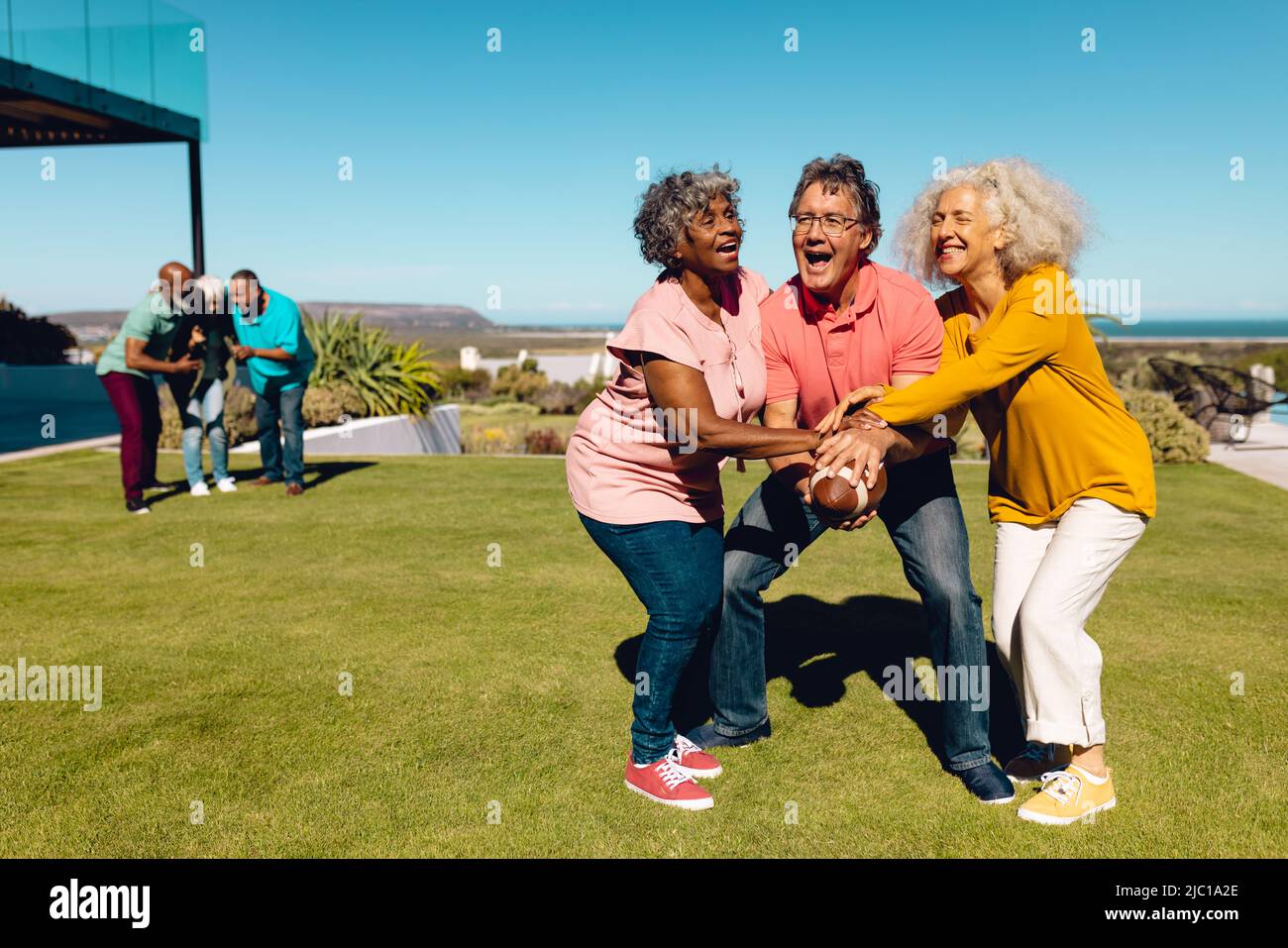 Donne senior multirazziale che bloccano l'uomo con la palla di rugby in cortile contro il cielo libero a casa pensionamento Foto Stock