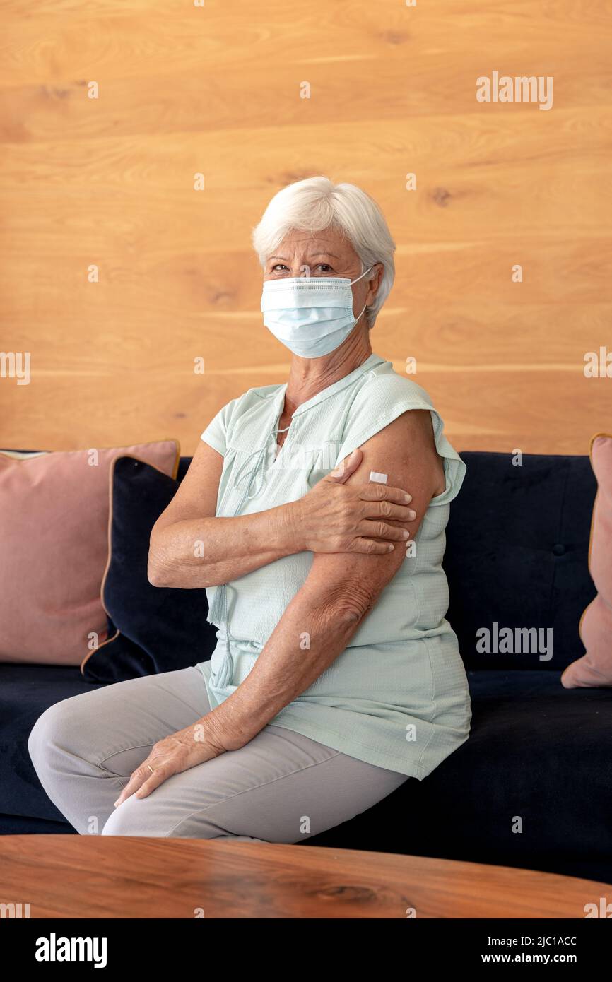 Ritratto della donna anziana caucasica che indossa maschera facciale che tiene il suo braccio con la fascia di aiuto seduta a casa Foto Stock