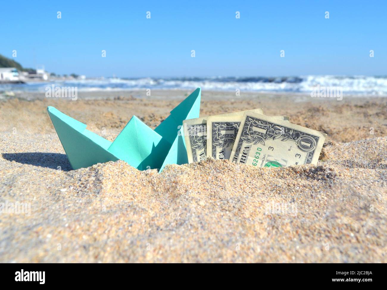 Piccola barca di carta blu e carta da tre dollari bollette mezzo in sabbia sulla spiaggia vicino al mare in estate giorno di sole. Concetto di viaggio, sogni, spese, denaro, finanza, turismo, riposo, vacanza obiettivi desidera Foto Stock