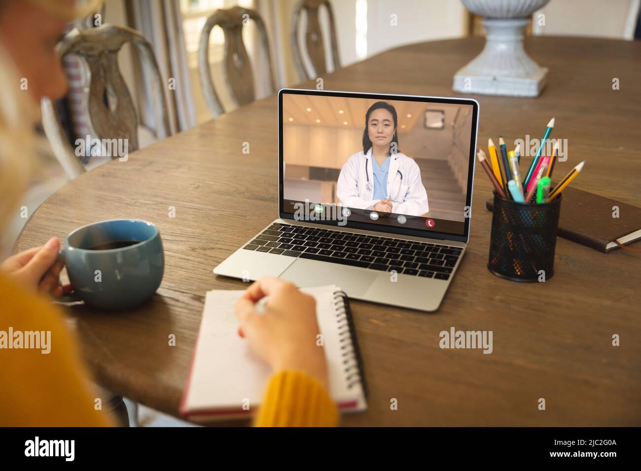 Medico asiatico in videochiamata parlando con donna d'affari caucasica scrivere sul libro alla reception Foto Stock