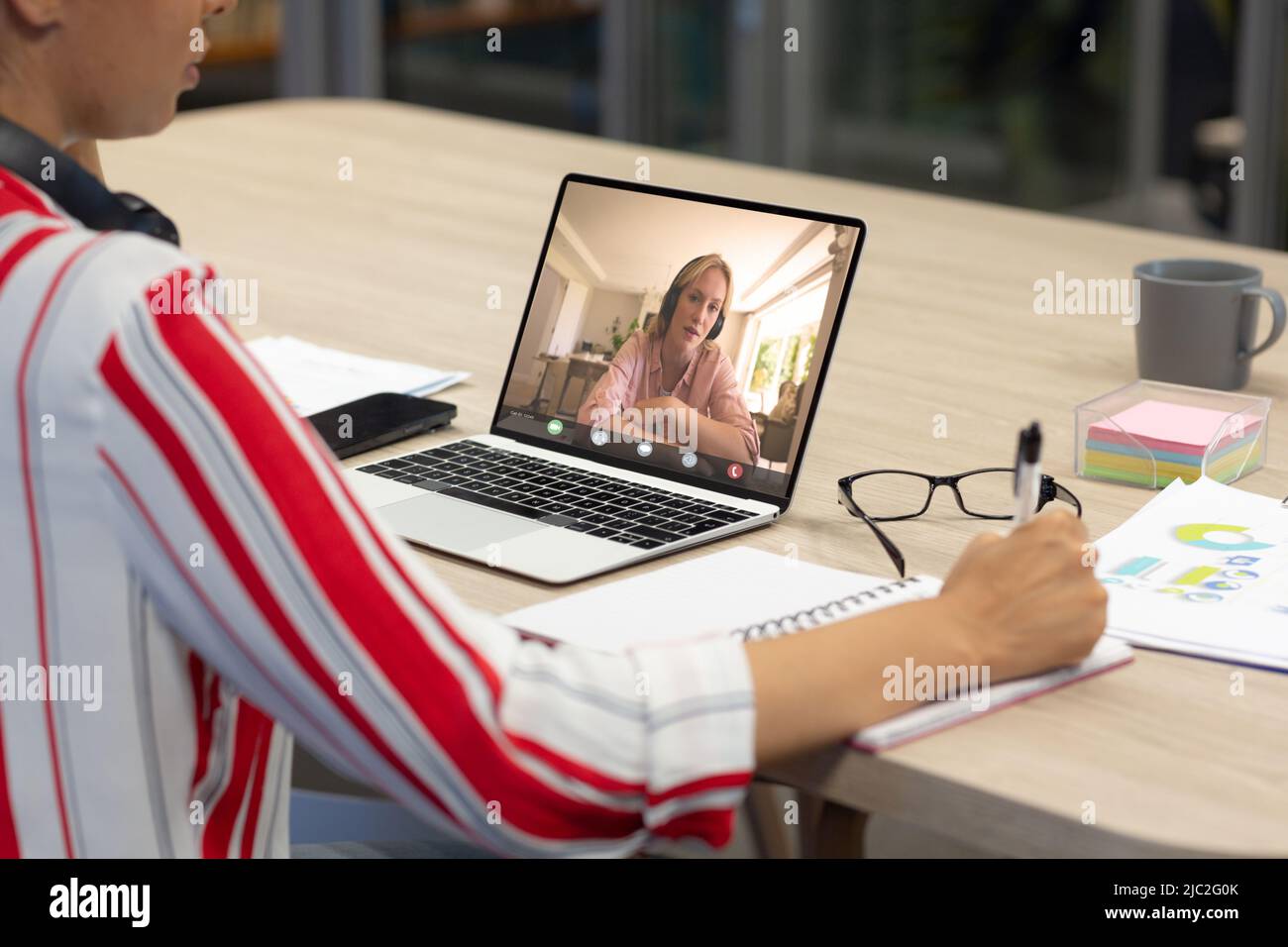 Donna d'affari caucasica in videochiamata con una collega asiatica che scrive sul diario alla scrivania in ufficio Foto Stock