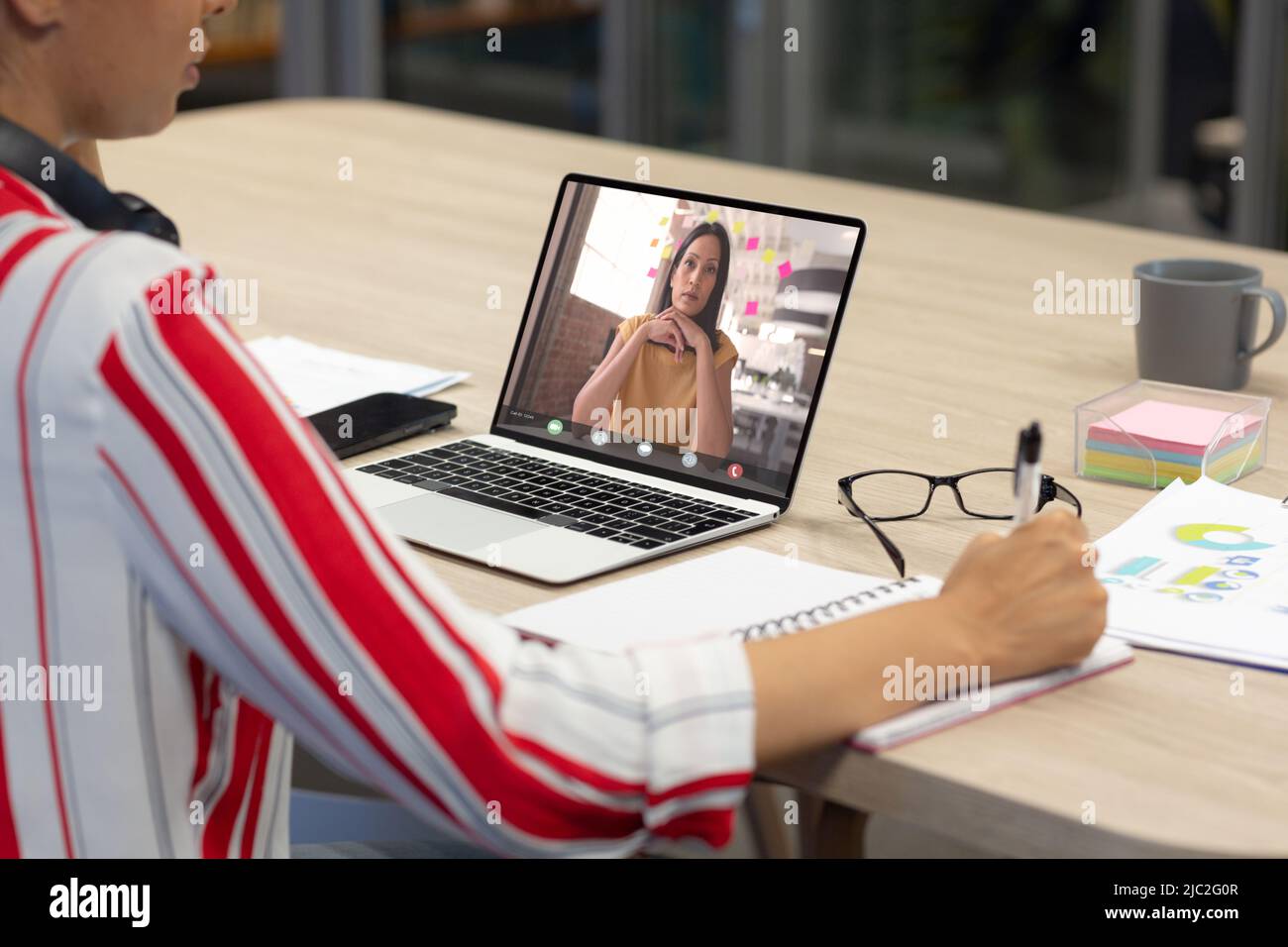 Donna d'affari asiatica in videochiamata con una collega asiatica che scrive sul diario alla scrivania in ufficio Foto Stock