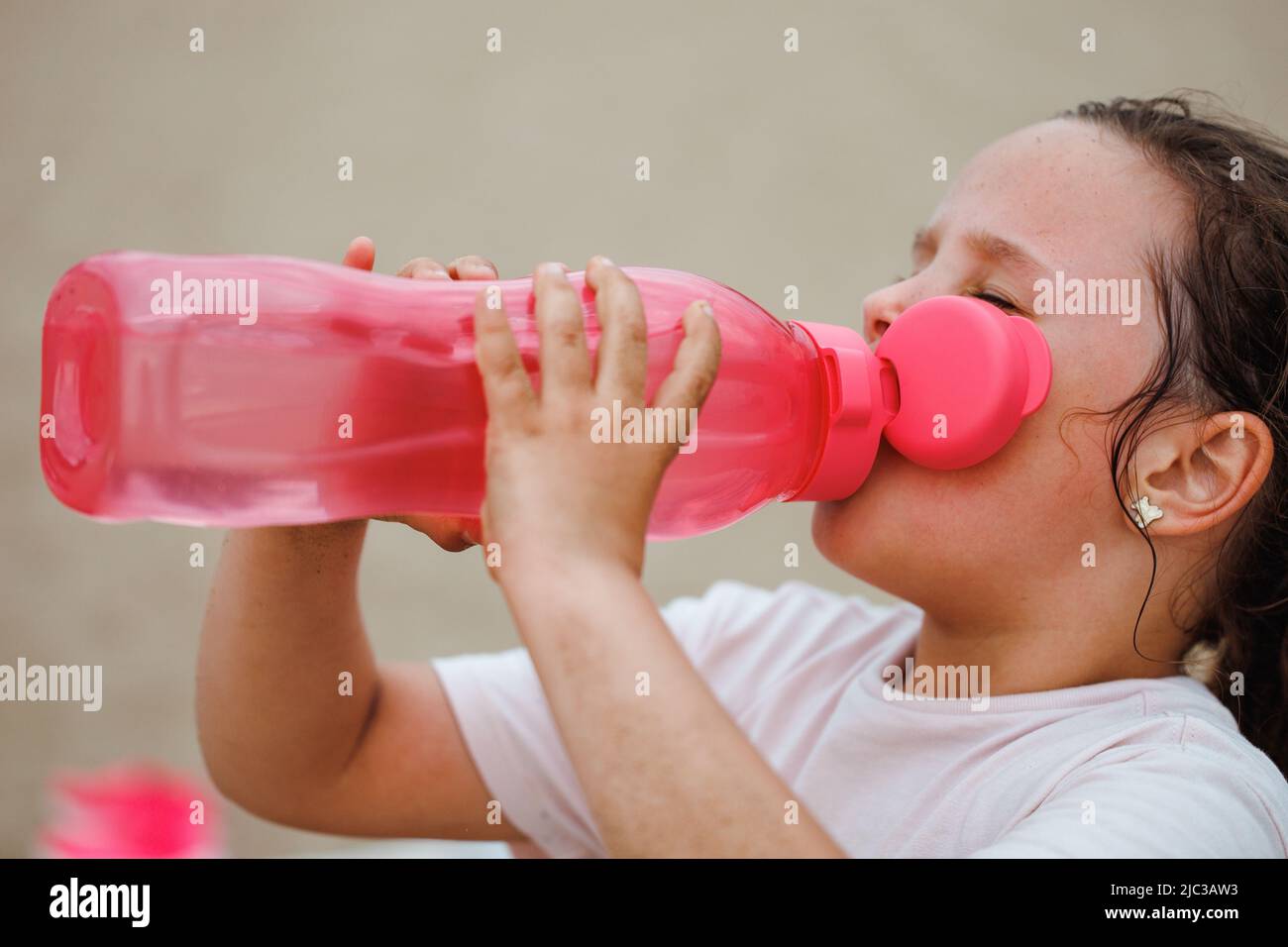 Vista laterale della bambina assetata con lunghi capelli scuri indossando una T-shirt bianca, acqua potabile da bottiglia di plastica rosa. Foto Stock