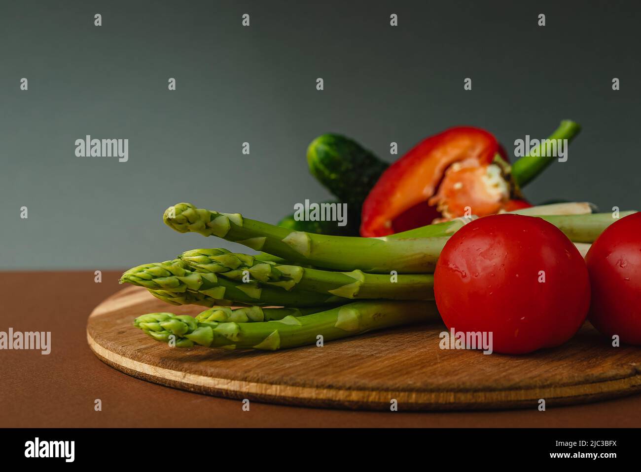 Le verdure si trovano su un asse di legno: Pomodori, asparagi, cetrioli, peperoni rossi. Marrone, sfondo grigio scuro. Luogo per il testo Foto Stock