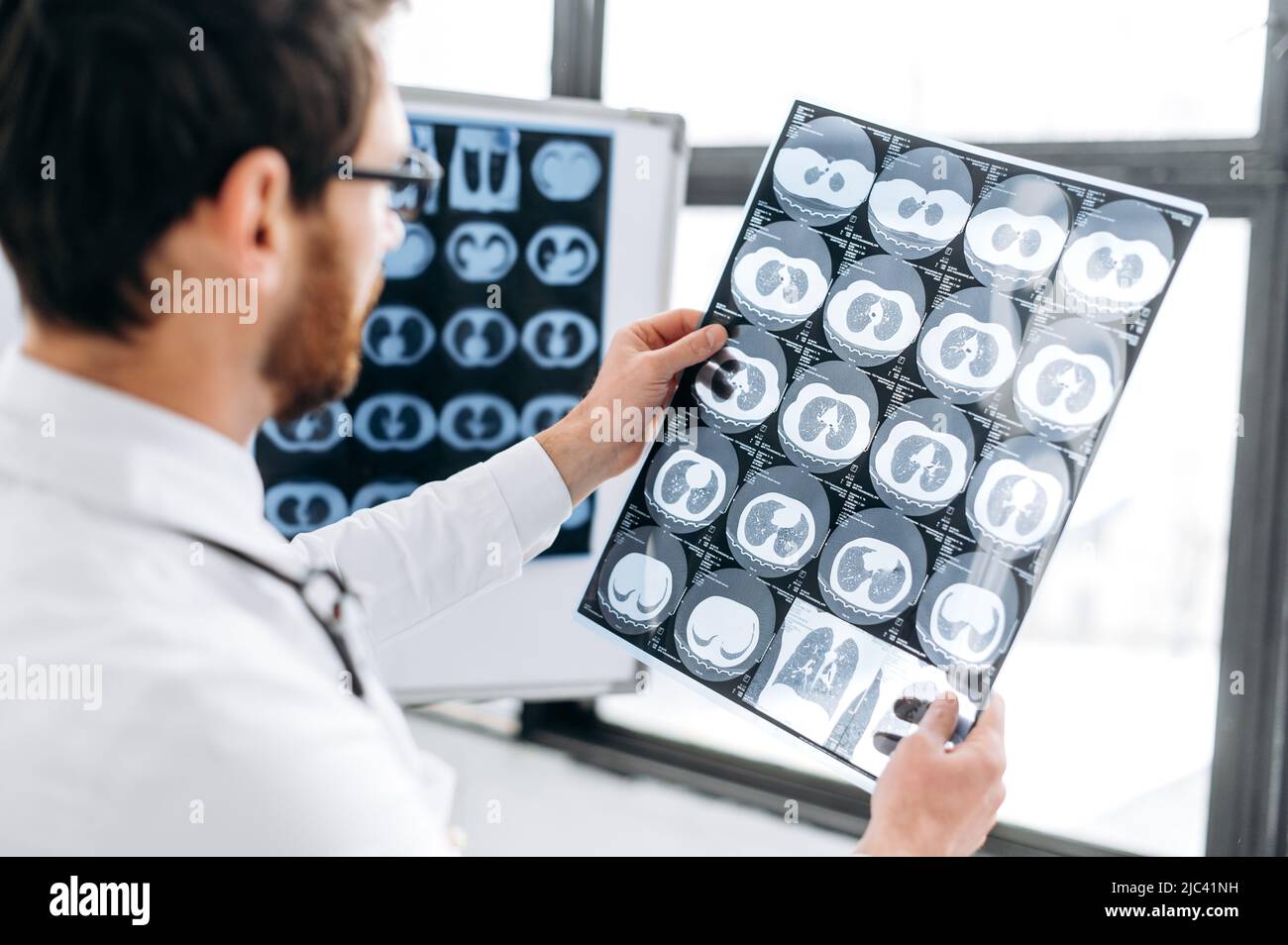 Studio radiologico. Medico intelligente focalizzato guardando la radiografia al suo gabinetto, terapista caucasico maschile in uniforme medica bianca e occhiali concentrato apprendimento radiografia del paziente, in piedi nel suo ufficio medico Foto Stock