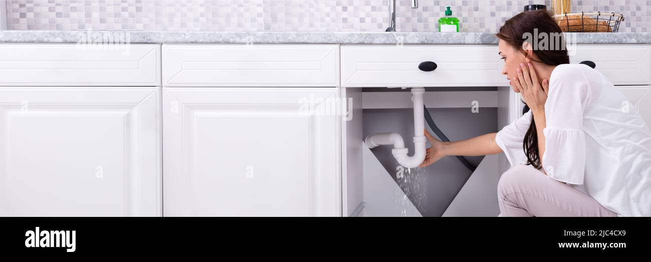 Vista posteriore di una donna che cerca di fermare la perdita di acqua dal tubo di lavello in cucina Foto Stock