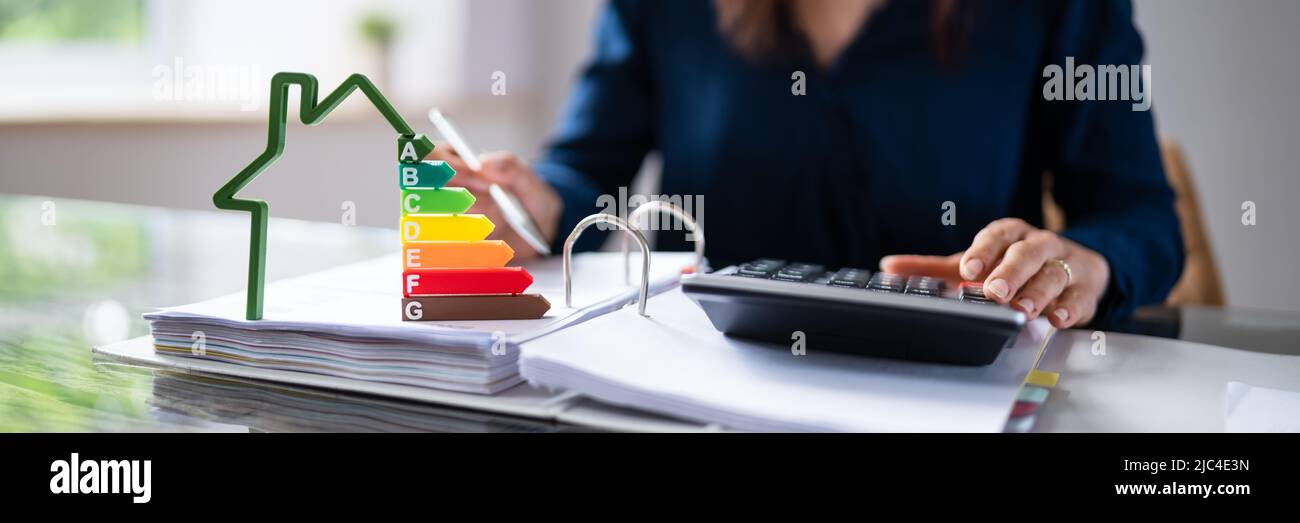 Close-up del modello di casa che mostra l'Efficienza Energetica tasso di fronte ai mediatori la mano utilizzando la calcolatrice Foto Stock