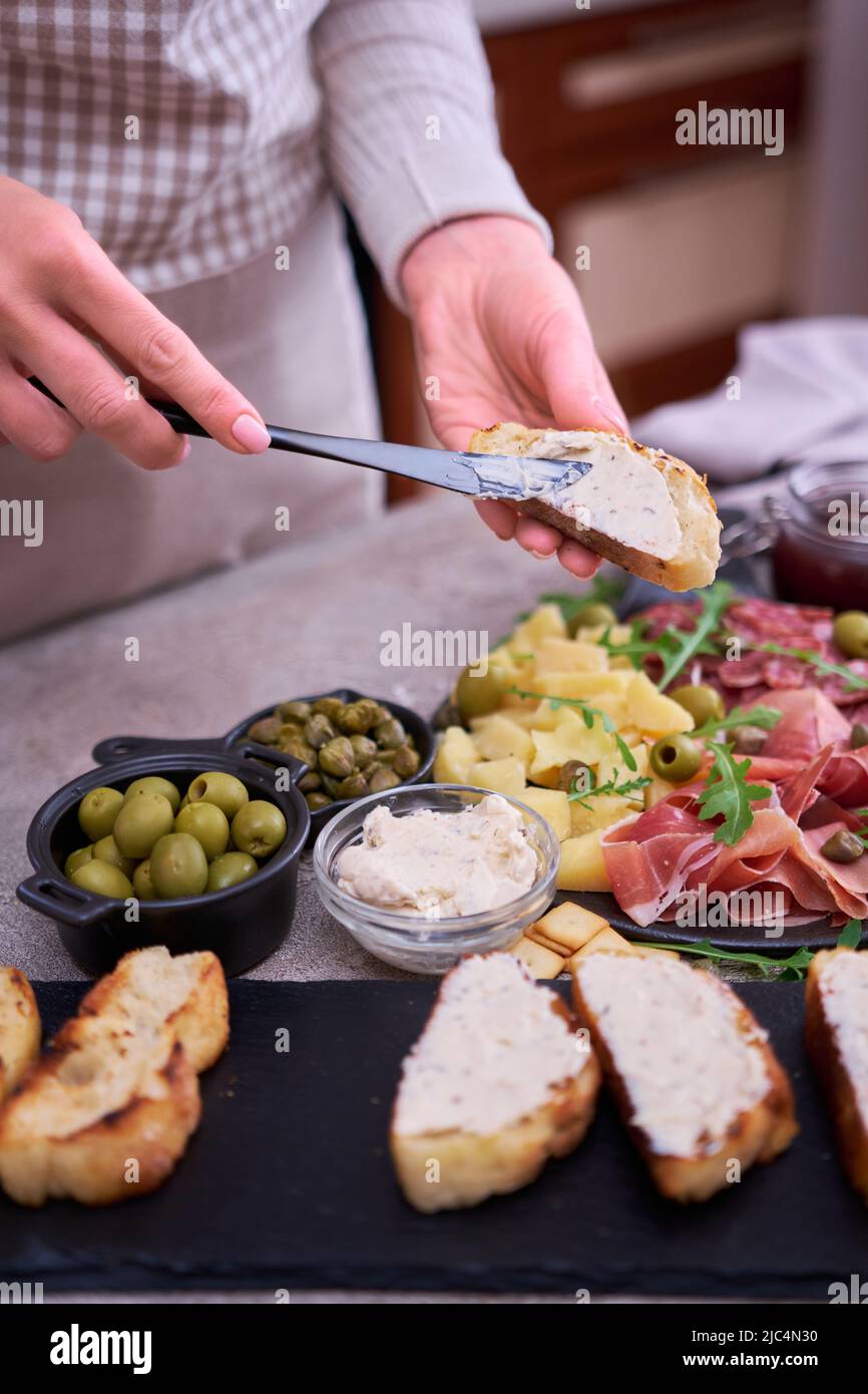 Donna che spalma il burro su un pezzo di baguette alla griglia con piatto di carne di antipasto italiano sullo sfondo Foto Stock
