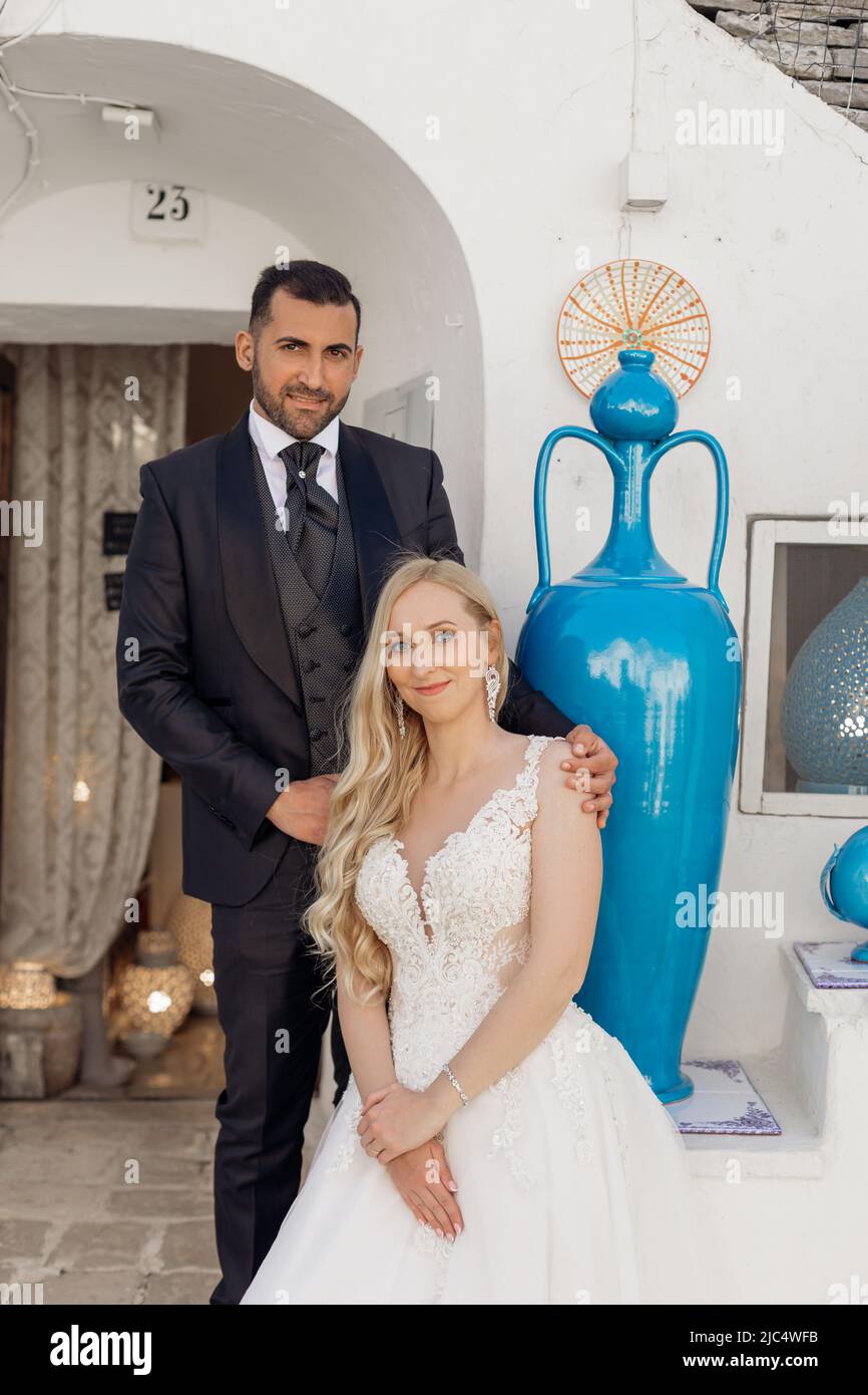 Ritratto di giovane sposa bionda in abito bianco e brunet sposo in abito sorridente e seduto vicino a casa vecchia città con antico vaso blu in Italia Foto Stock