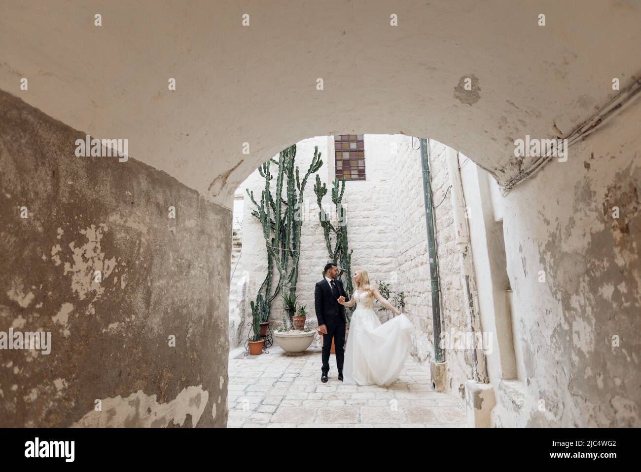 Ritratto all'interno di arco in cemento di giovane sposa bionda in abito bianco e brunet sposo in vestito posa vicino al vecchio edificio della città in Italia, sfondo urbano Foto Stock
