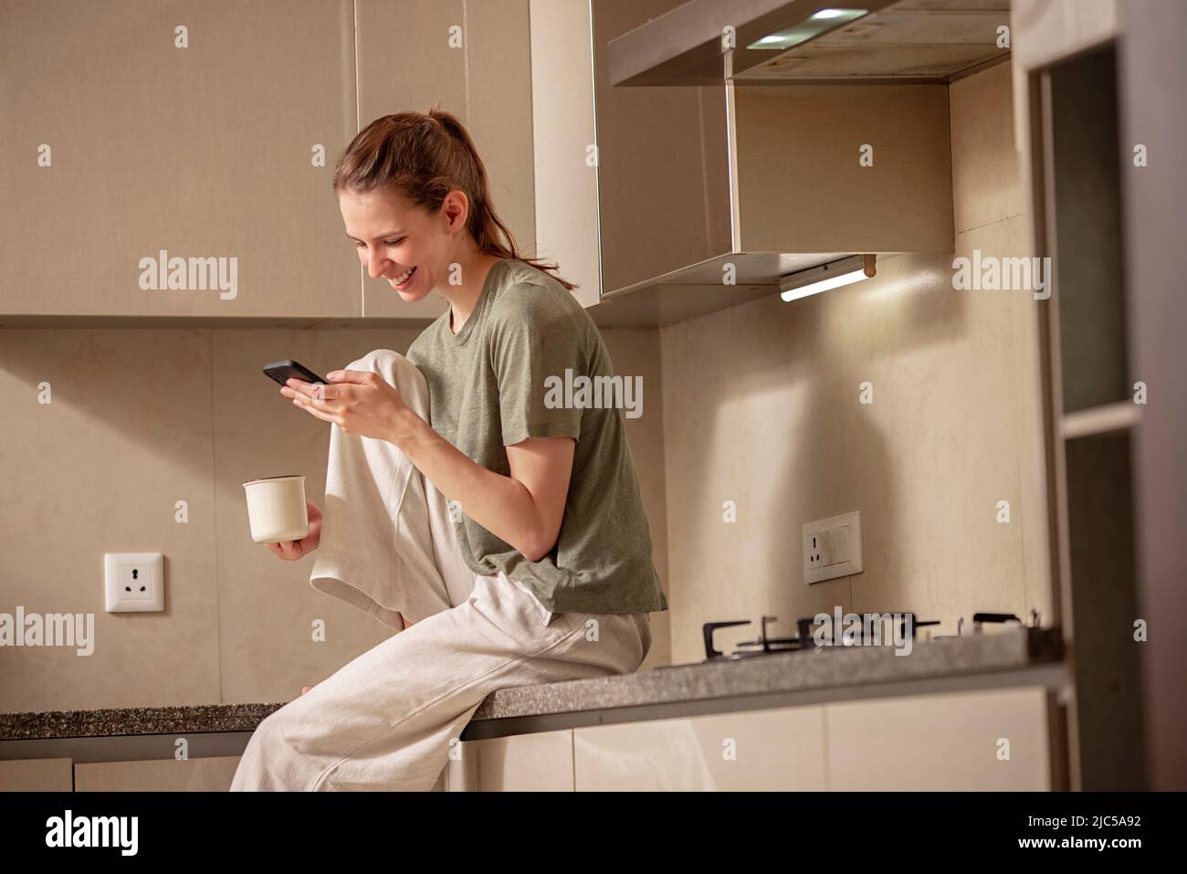 Giovane donna seduta in cucina e con un telefono cellulare Foto Stock