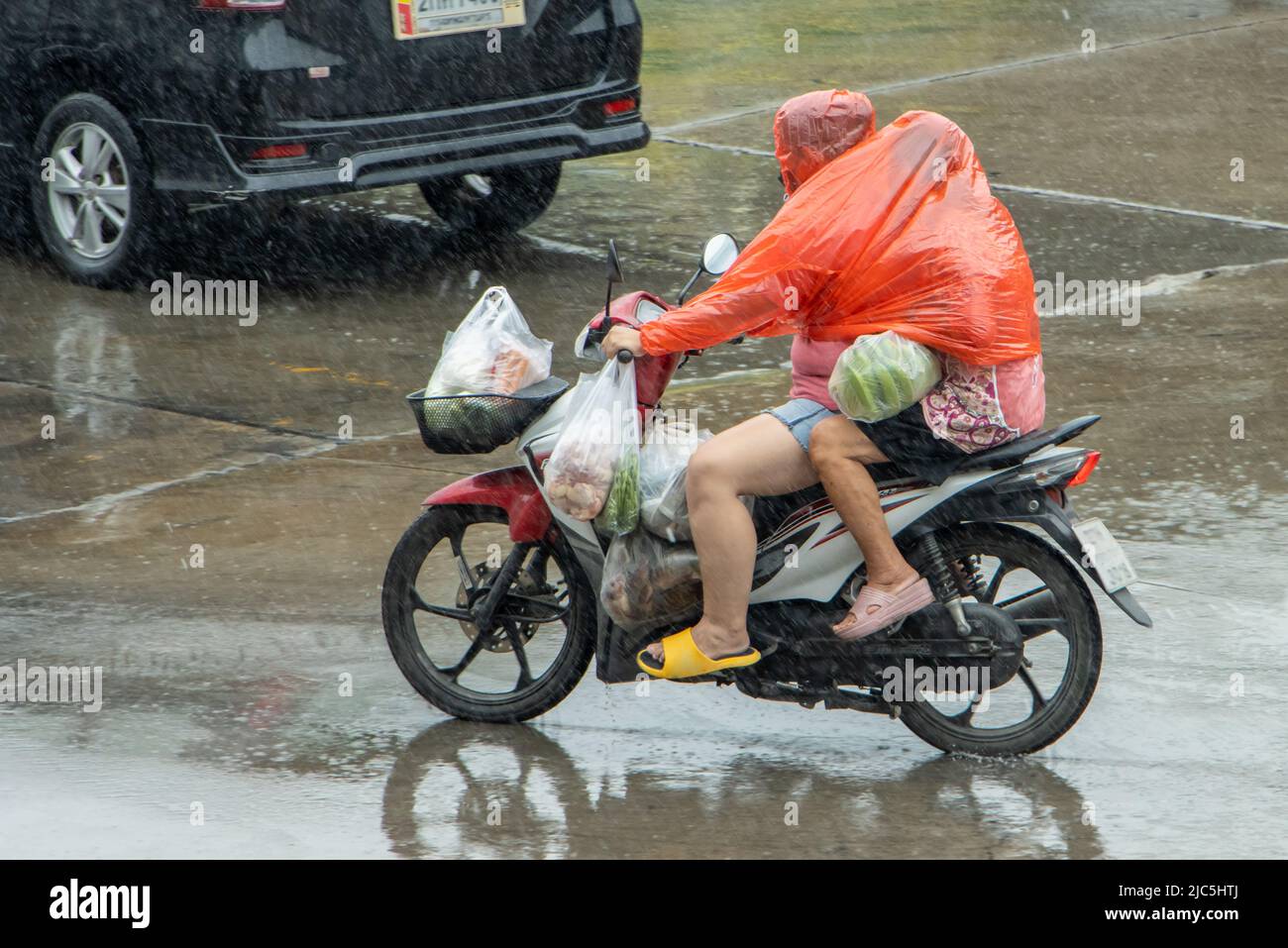 Una coppia in un impermeabile con sacchetti di plastica del cibo cavalcano una motocicletta sotto la pioggia Foto Stock