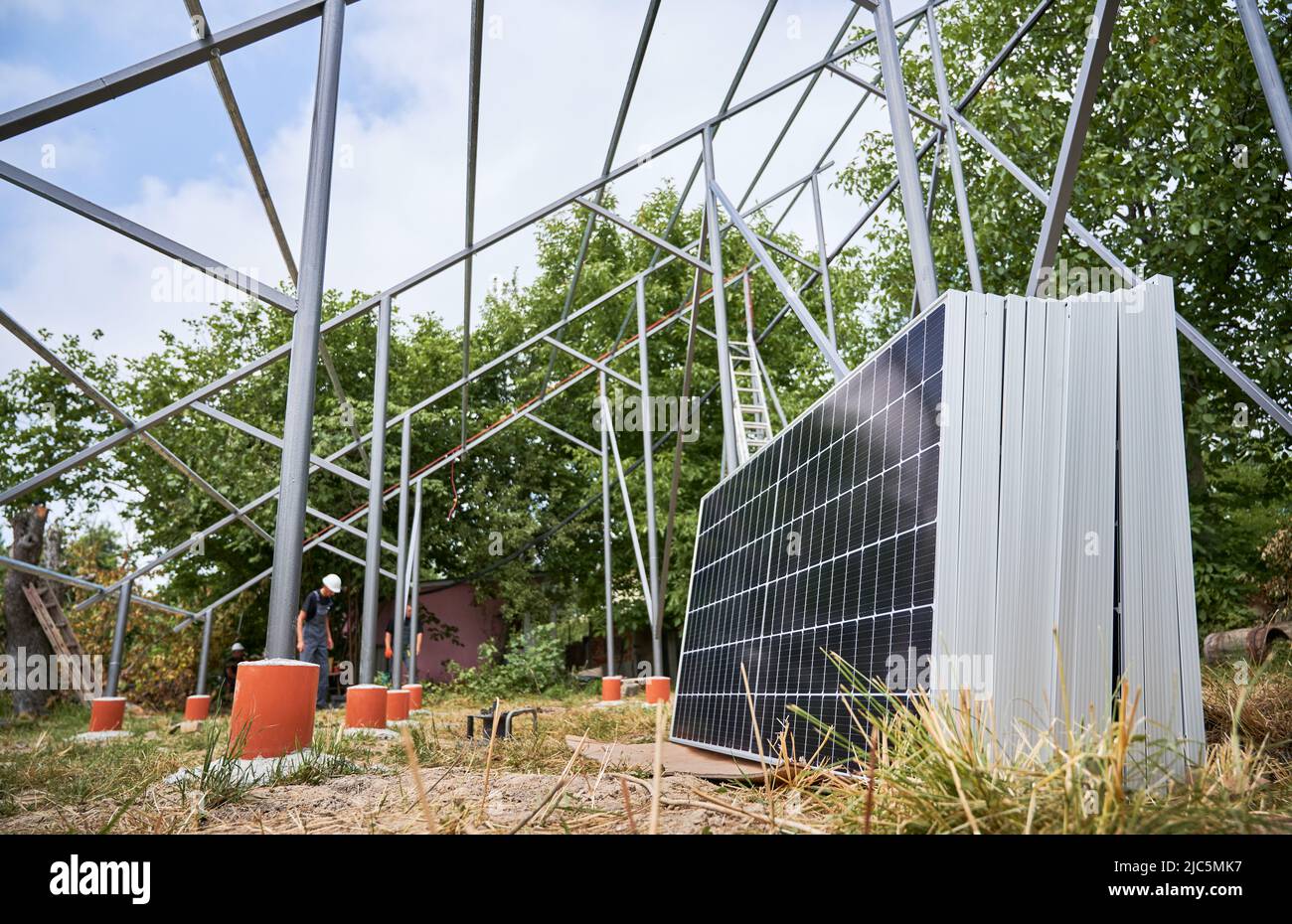 Strutture di supporto in metallo, moduli solari per la generazione di energia elettrica attraverso l'effetto fotovoltaico e lavoratori maschi. Installatori solari che assemblano pannelli solari in aree erbose con alberi. Foto Stock