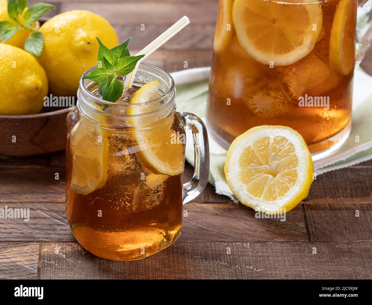 Bicchiere di tè freddo con fette di limone, menta e ghiaccio su un rustico tavolo di legno. Caraffa e limoni sullo sfondo Foto Stock