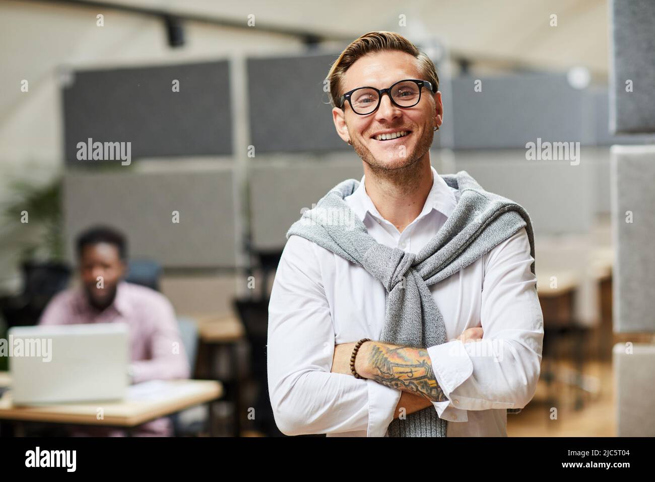 Ritratto di un giovane sviluppatore di app entusiasta con maglione avvolto intorno al collo in piedi con braccia incrociate in un ufficio moderno Foto Stock