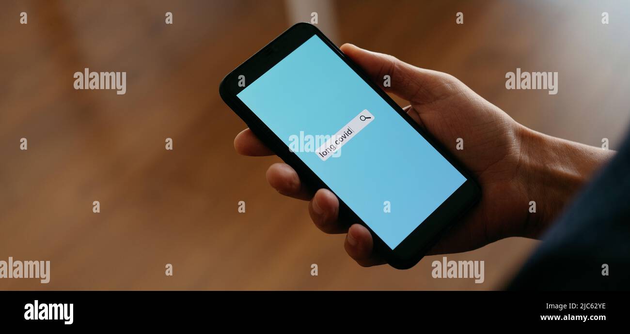 un uomo al chiuso sta cercando il testo lungo covid in un motore di ricerca internet simulato nel suo smartphone, in un formato panoramico da utilizzare come banner web o. Foto Stock