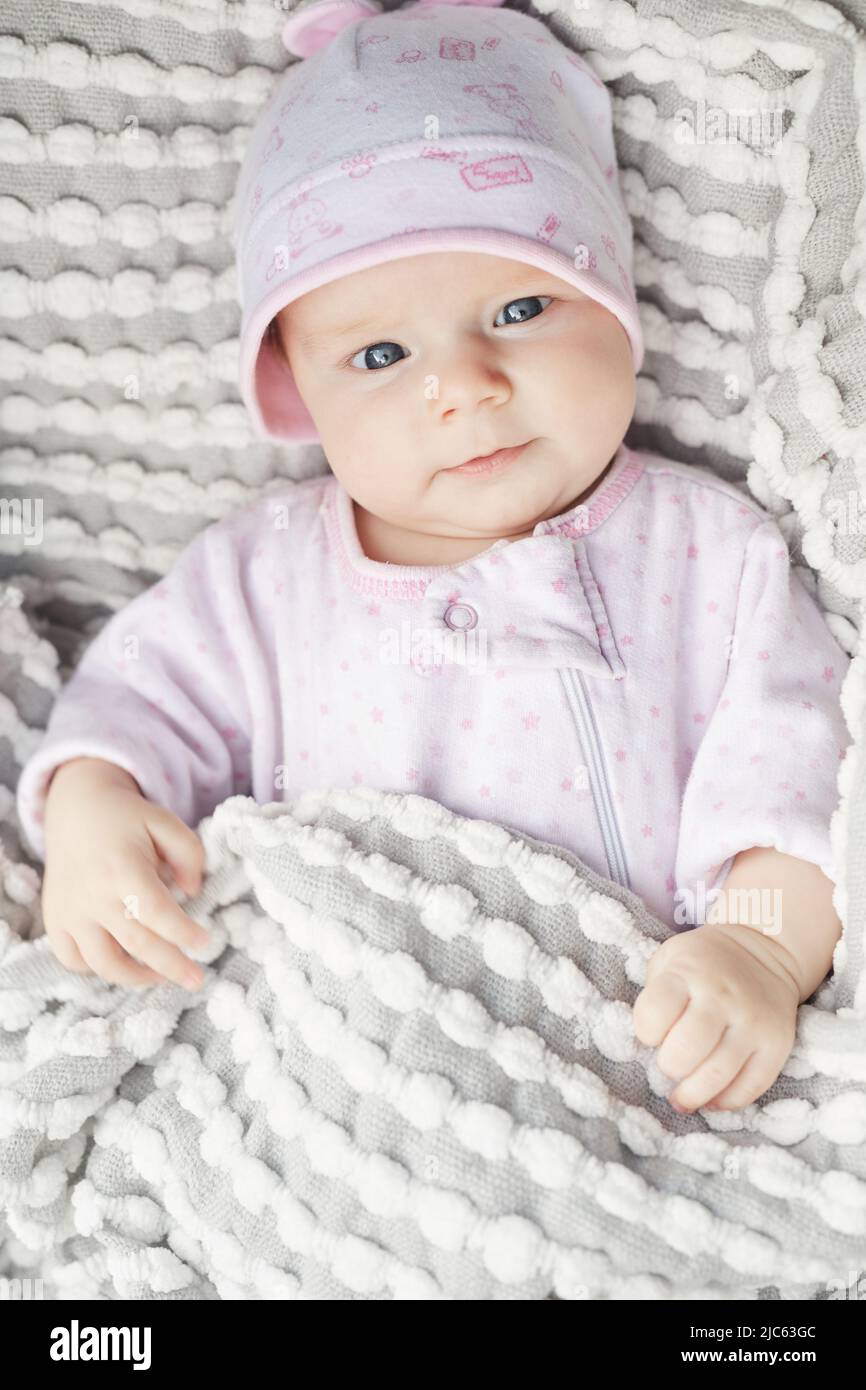 Bambino dolce e piccolo con grandi occhi blu in costume bianco e rosa e cappello sdraiato sotto una comoda coperta grigia Foto Stock
