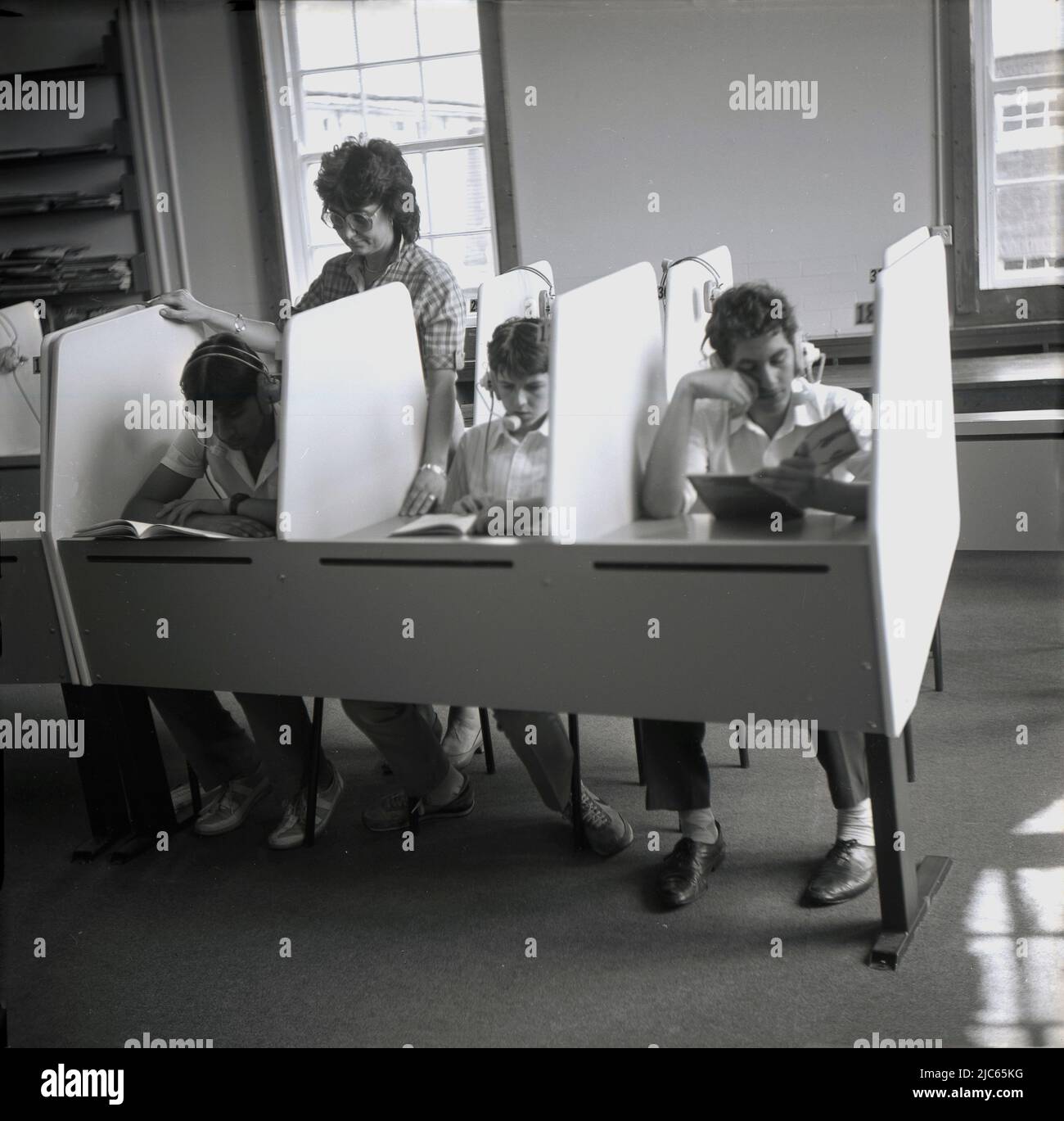 1970s, storici, studenti di lingua seduti in una scrivania con lati in pannellino, con cuffie su. Una donna insegnante in piedi, che sovrintendere i ragazzi adolescenti Foto Stock
