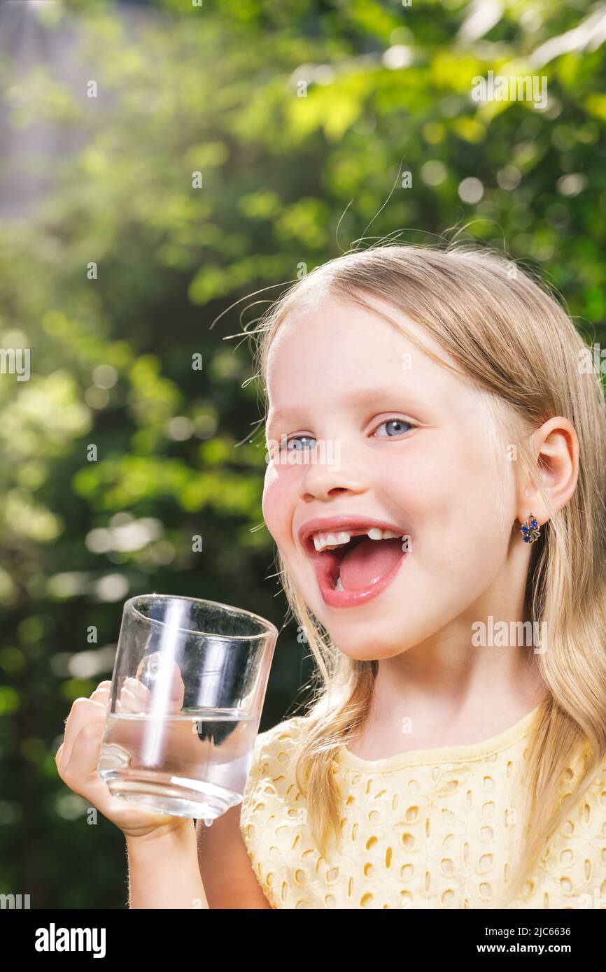 Ragazza piccola felice che indossa abito giallo che tiene bicchiere d'acqua in un giardino estivo - stile di vita sano concetto Foto Stock