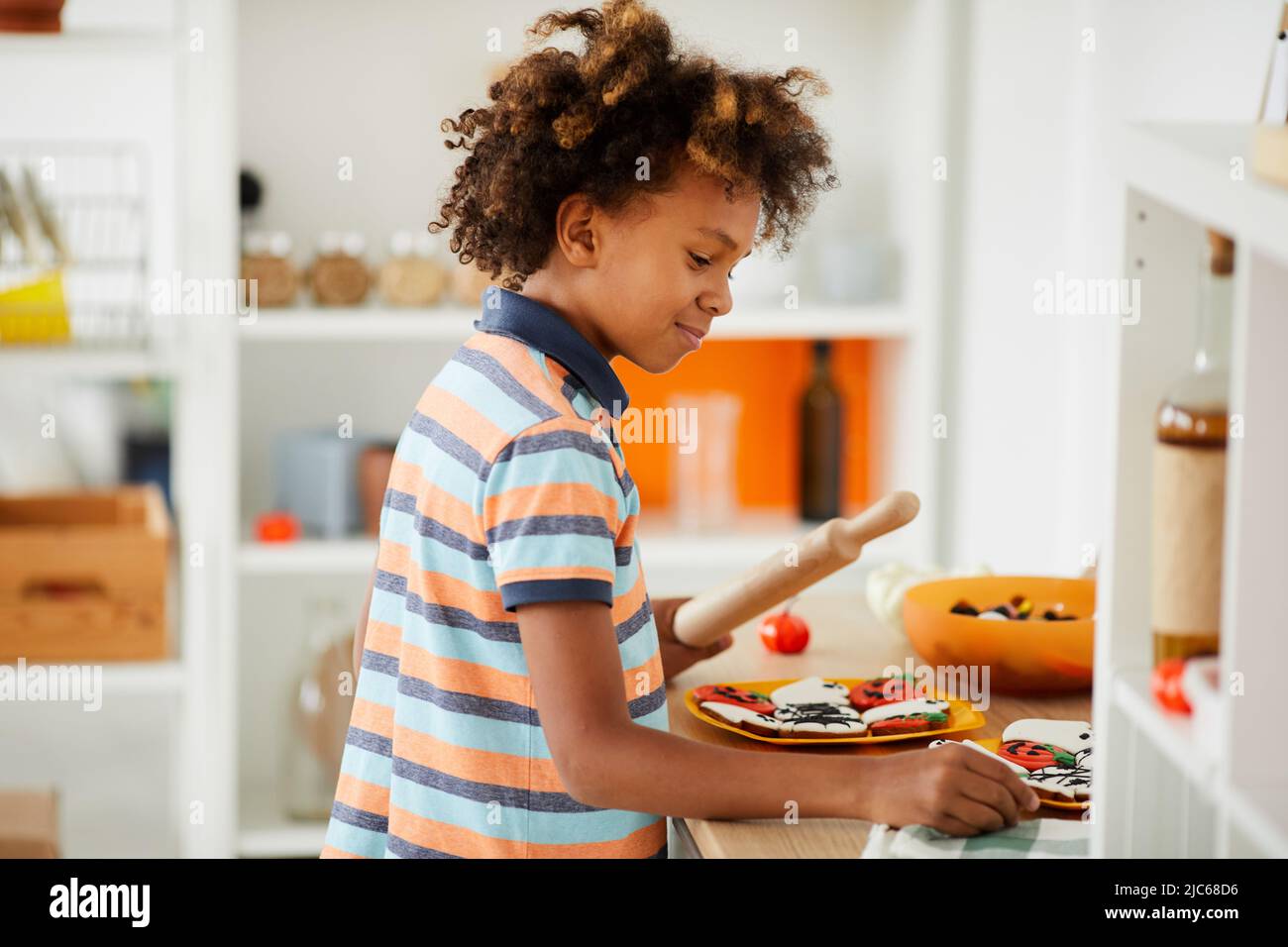 Sorridente ragazzo afroamericano in tshirt colorata in piedi al banco della cucina e utilizzando il rolling pin mentre cucinano biscotti di pan di zenzero Foto Stock