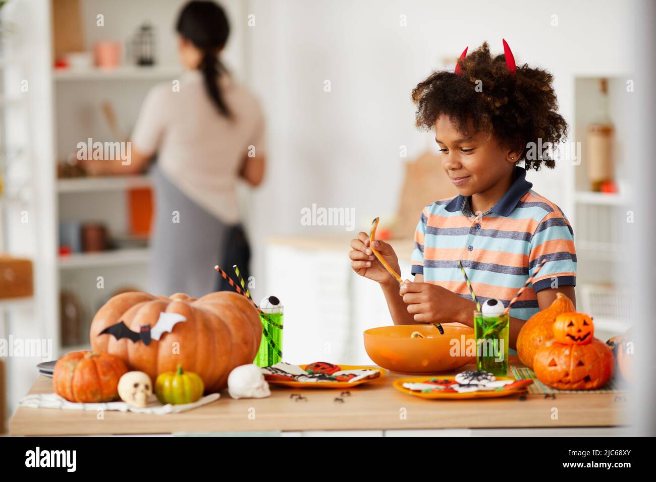 Sorridendo ragazzo nero carino con corna diavoli in piedi al banco con varie caramelle di Halloween e mangiare verme di marmellata Foto Stock