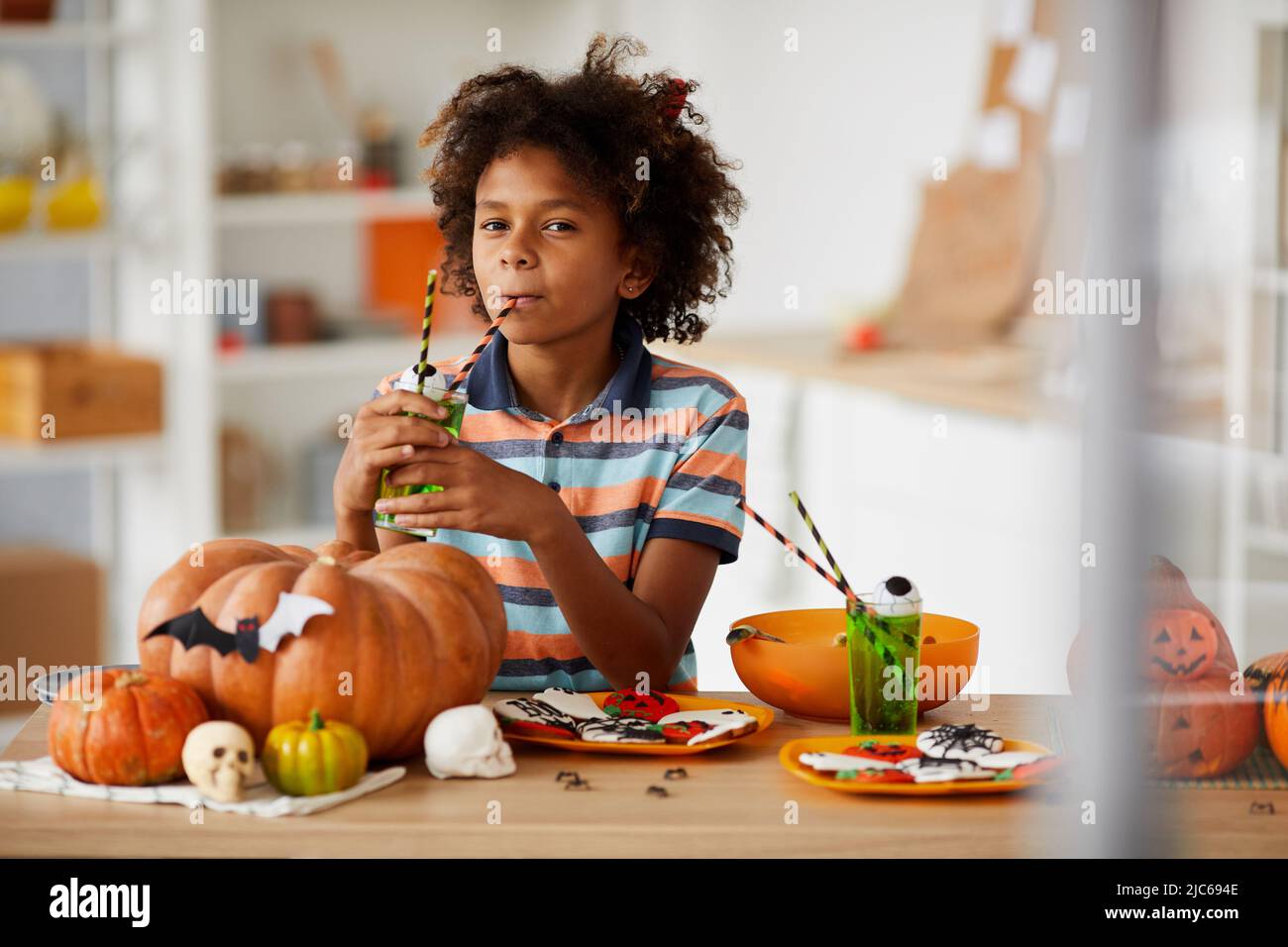 Pensive Black Boy con capelli ricci appoggiati su un bancone di legno con dolci e succo di frutta da paglia Foto Stock