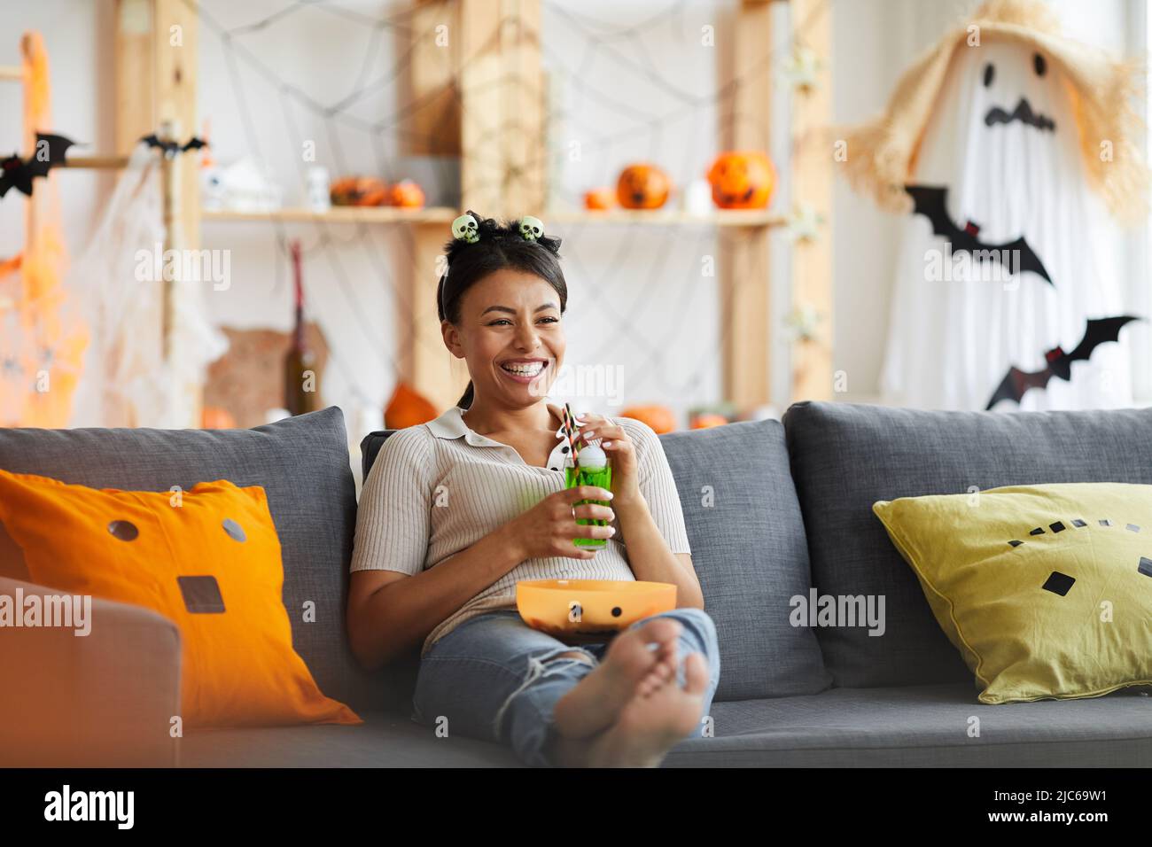 Positiva donna nera rilassata in fascia con teschi giocattolo seduti sul divano e bere limonata mentre si guarda la tv su Halloween Foto Stock