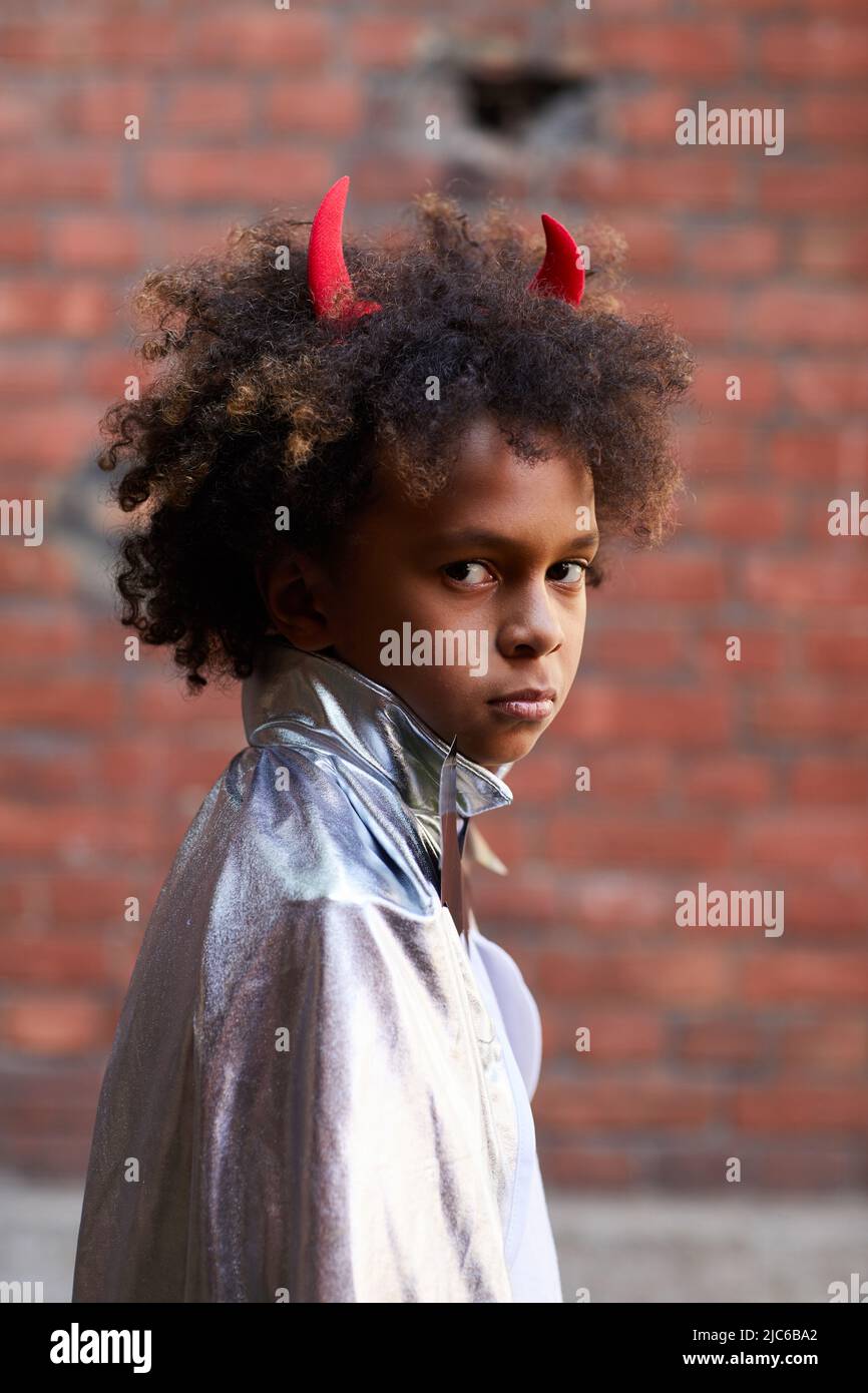 Ritratto di grave ragazzo diavolo arrabbiato di etnia africana indossando corna rosse e mantello d'argento in piedi fuori Foto Stock