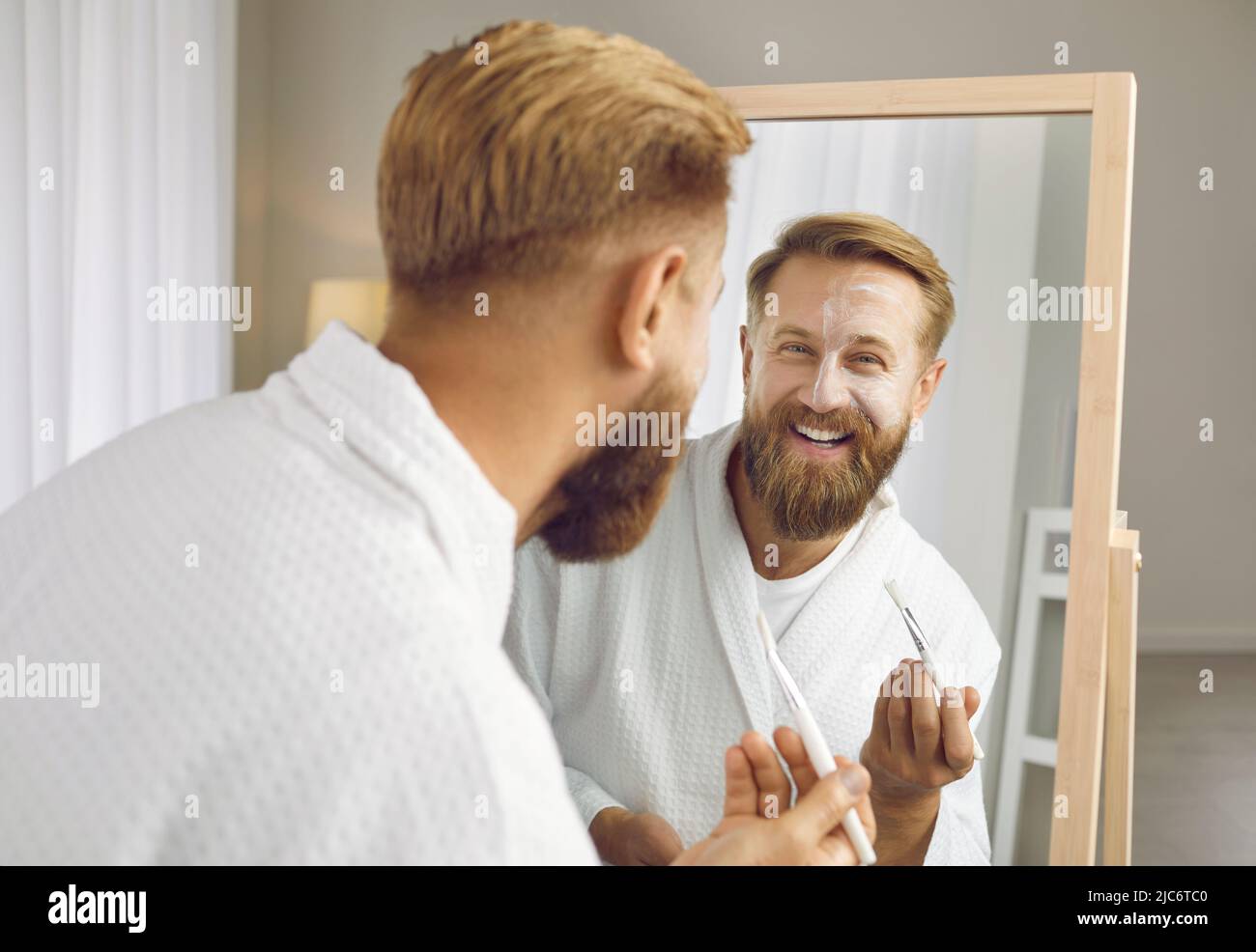 Felice bell'uomo che guarda nello specchio, applicando una maschera di argilla bianca e sorridendo Foto Stock