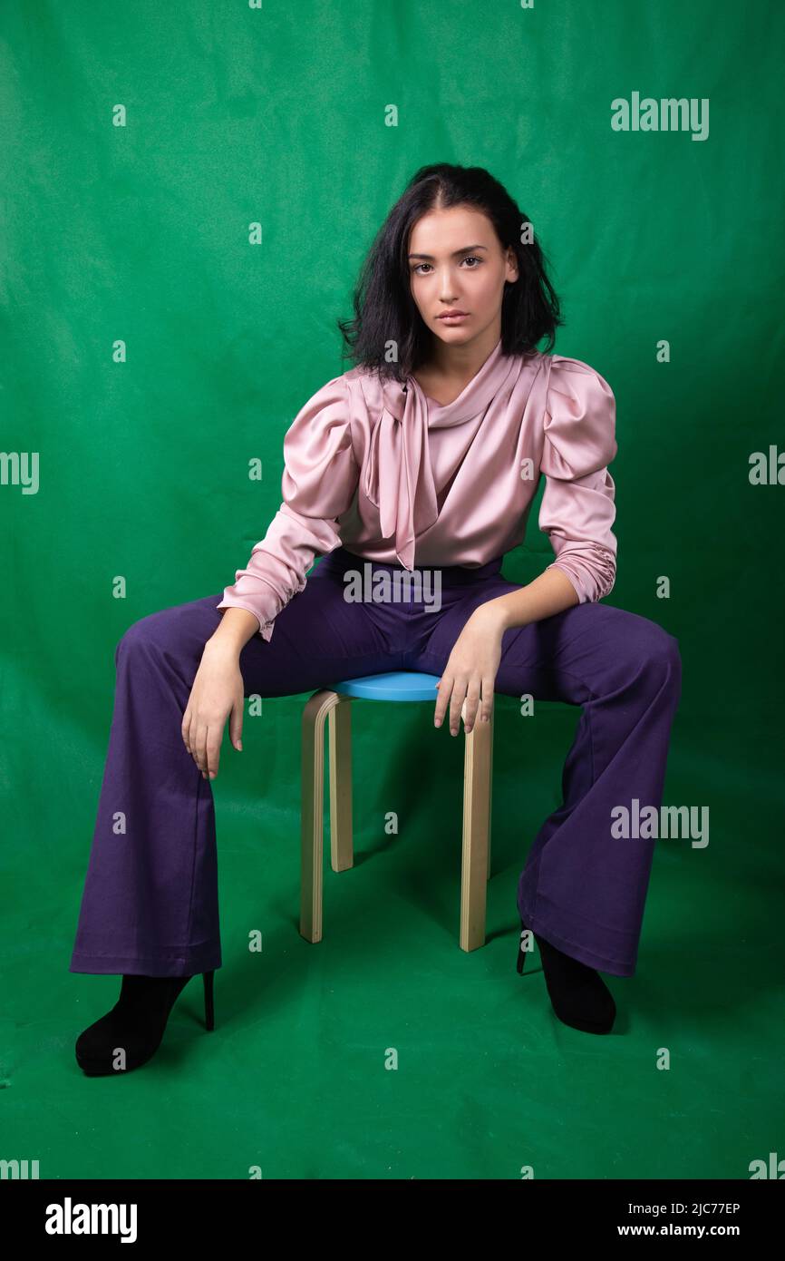 Giovane grazioso modello seduto su uno sgabello blu poggiando i gomiti sulle ginocchia in studio Foto Stock