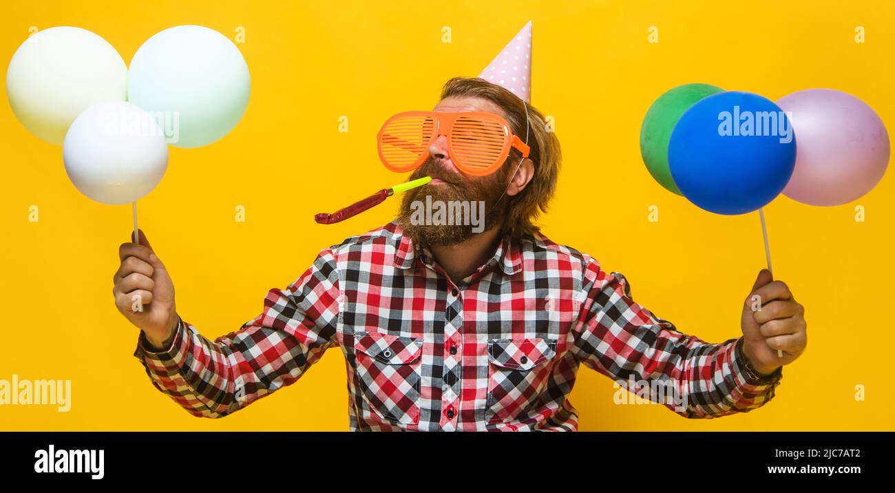 Uomo bearded in cappello di compleanno e occhiali divertenti con palloncini che soffiano noisemaker. Ora di festa. Vacanza. Foto Stock