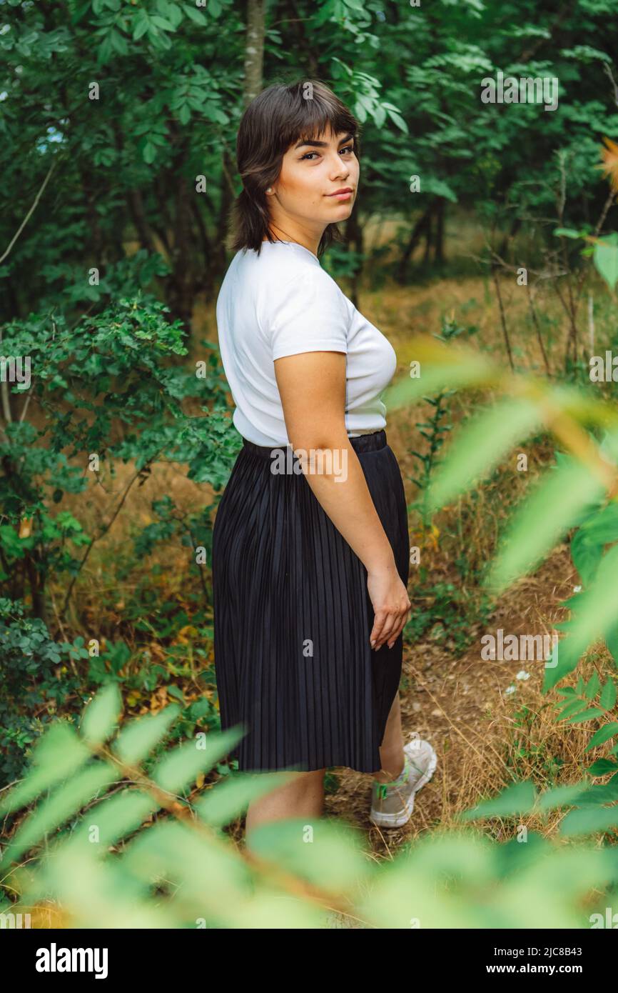 Una giovane ragazza con corti capelli neri, una T-shirt bianca e una gonna nera nel bosco tra alberi verdi e erba verde Foto Stock