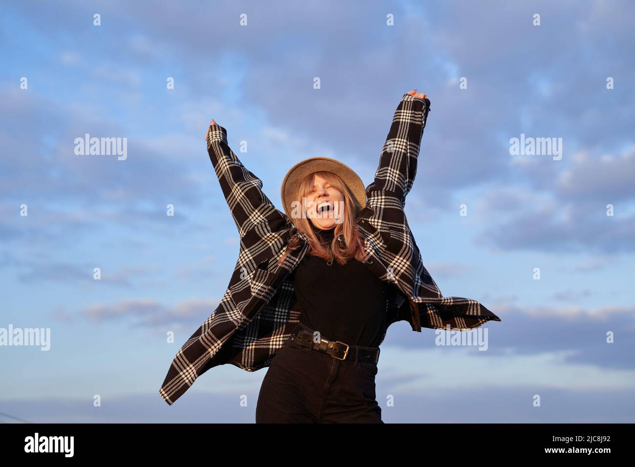 Giovane ragazza eccitata hipster in cappello, jeans neri e maglietta che ballano sentendosi gioiosa o spensierata emozione di fronte al cielo blu nuvoloso. Concetto trionfo, vincente, espressivo o positivo. Immagine di alta qualità Foto Stock
