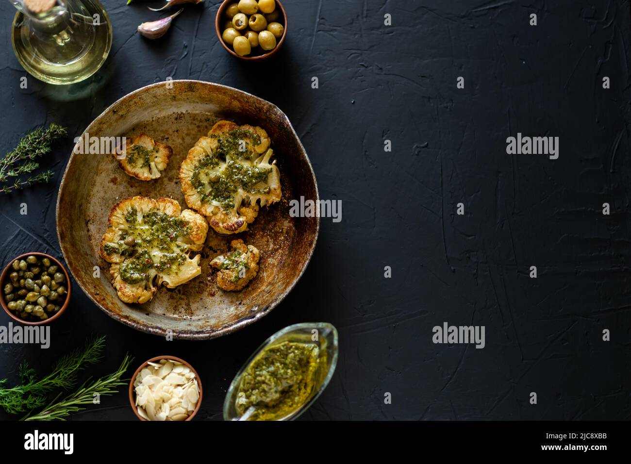 La bistecca di cavolfiore con spezie si trova in una padella. Olio d'oliva, salsa di chimichurri, erbe aromatiche, varie spezie affiancate. Sfondo scuro. Posiziona per testo. Foto Stock