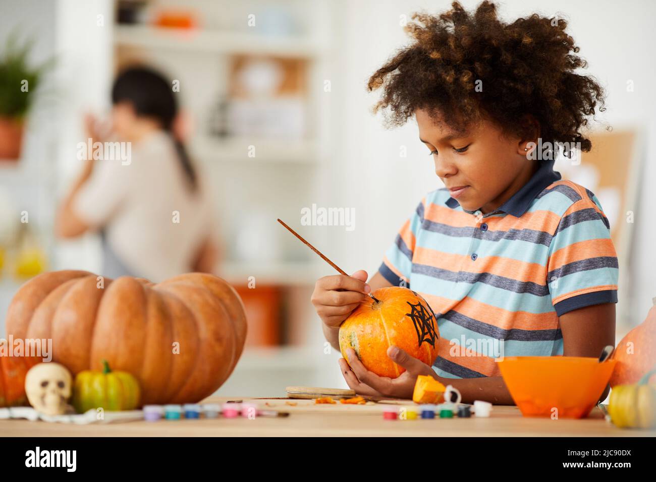 Sorridente bambino creativo con acconciatura Afro appoggiata sul banco con gouaches e fare il disegno sulla zucca Foto Stock