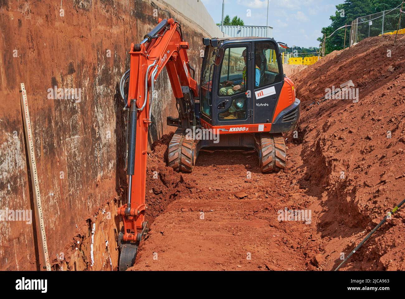 Worker utilizza un escavatore per scavare una trincea sotto e strada accanto a un muro di contenimento in cemento armato Foto Stock