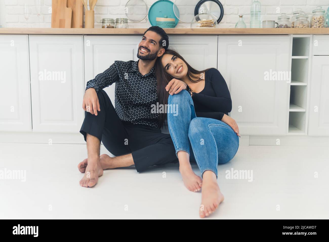 Gioioso multirazziale elegante coppia seduta a piedi nudi sul pavimento, appoggiata su mobili da cucina bianchi. Affetto e concetto di amore. Foto di alta qualità Foto Stock