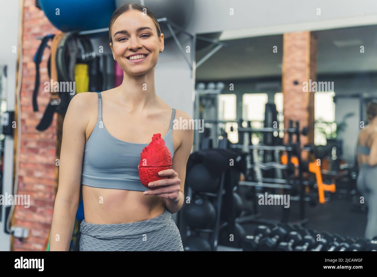 Giovane donna bianca ed eccitata in una palestra ben attrezzata, sorridente alla macchina fotografica, con un cuore umano falso rosso. Foto di alta qualità Foto Stock