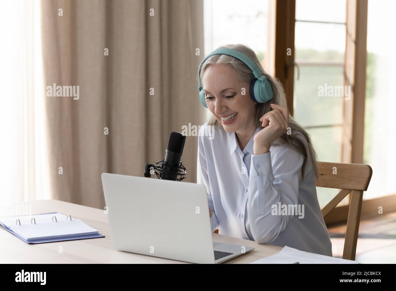 La donna di mezza età si siede alla scrivania con un computer portatile e parla nel microfono Foto Stock