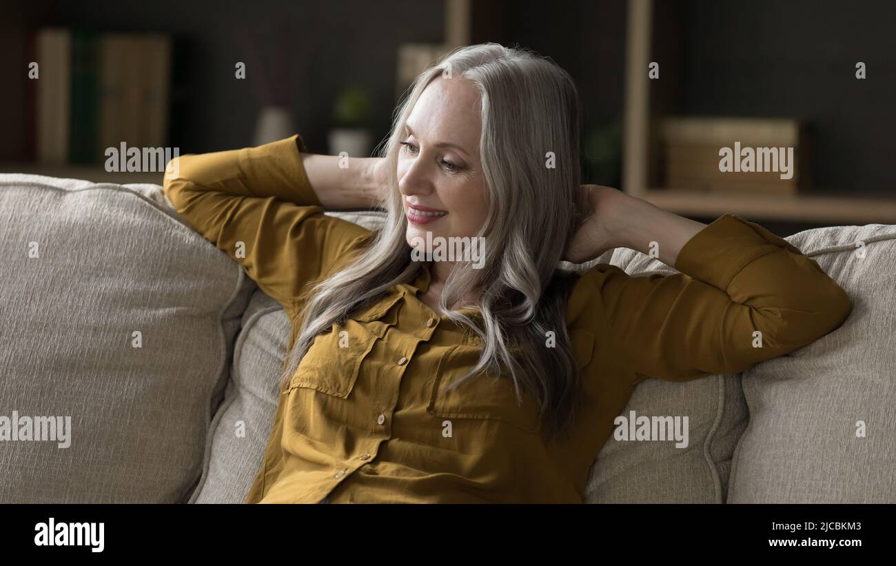 Donna di mezza età con lunghi capelli grigi seduta su un comodo divano Foto Stock