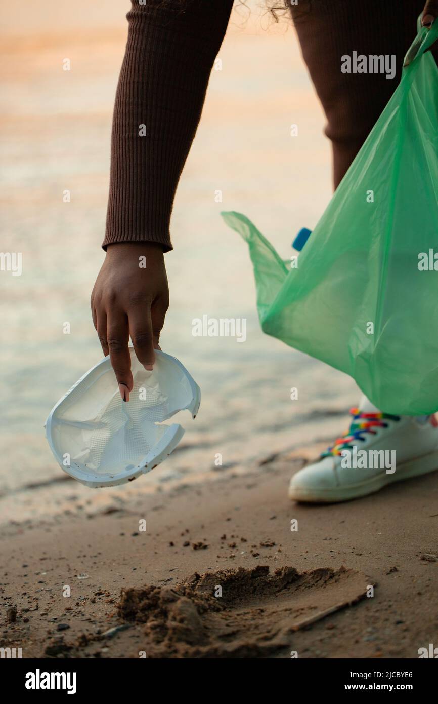 Foto ritagliata della donna africana che raccoglie spazzatura versato dalla sabbia sulla spiaggia in sacchetto di plastica verde. Ecologia, inquinamento. Foto Stock