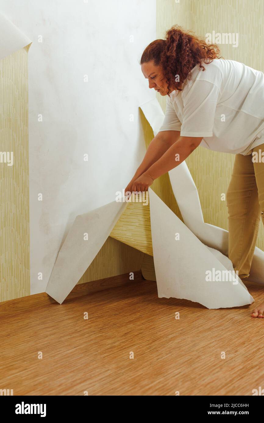 Vista laterale della giovane donna in rilievo che indossa una T-shirt bianca, pantaloni beige, appoggiati per strappare la vecchia carta da parati dal muro. Foto Stock