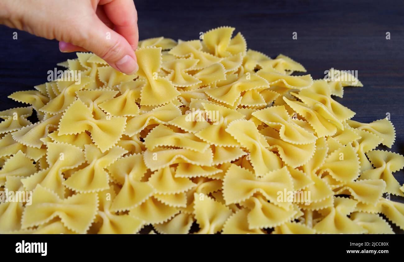 A mano che sceglie un pezzo di Farfalle o Pasta di Bow-tie dal suo pile su sfondo di legno nero Foto Stock