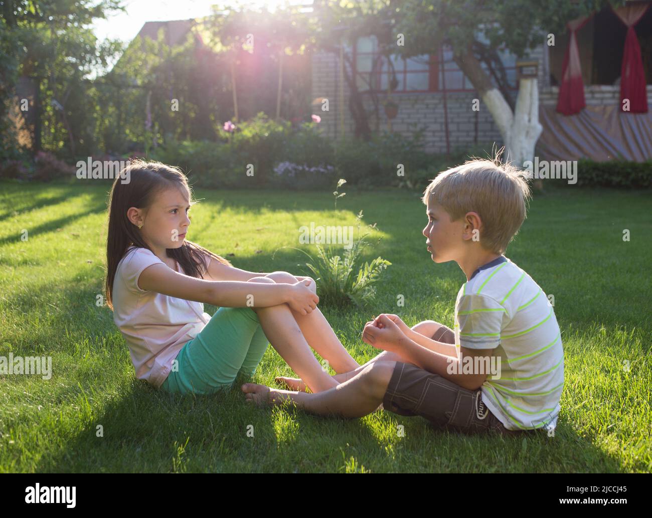 In estate, ragazzo e ragazza di 6-7 anni siedono sull'erba nel cortile del sole che tramonta in giardino, parlando e giocando nel verde del parco soleggiato. Frien Foto Stock