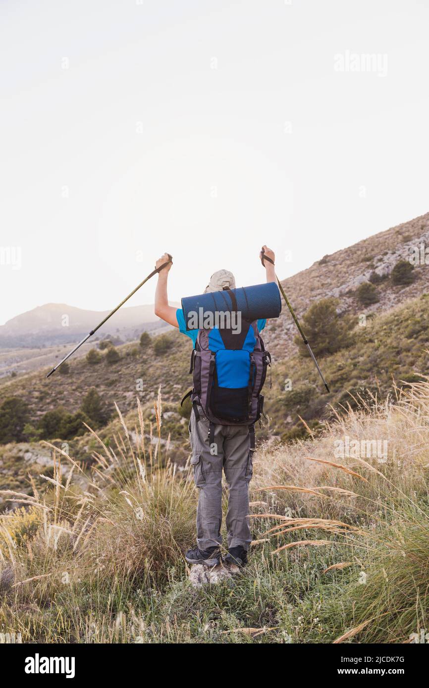 Colpo verticale di un escursionista, adulto, maschio, caucasico, con uno zaino, stuoia, pali, vestito di t-shirt blu, cappuccio e pantaloni grigi è sulla montagna garzando Foto Stock